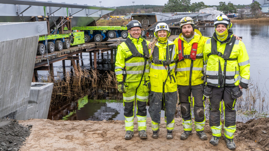 Fire fornøyde og smilende medarbeidere står tett sammen foran brokonstruksjon over vannet i Karlstad, Sverige.